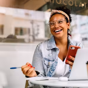 Mulher preta sorridente sentada de frente para um computador com uma caneta e um celular em mãos.