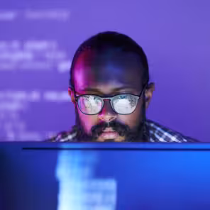 Programador de óculos trabalha em frente a computador em luzes roxas.
